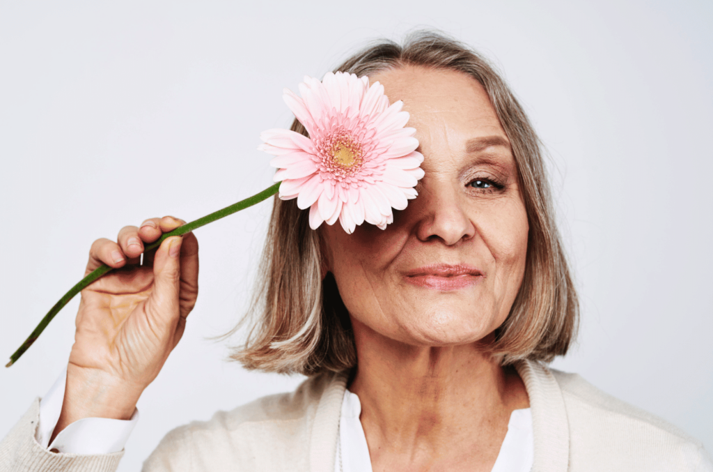 Femme mature souriante tenant une fleur rose devant son œil, représentant la féminité et le bien-être naturel à travers un accompagnement personnalisé pour la ménopause en naturopathie avec Agnès Kerguillec, Naturopathe Lyon Vienne et Visio